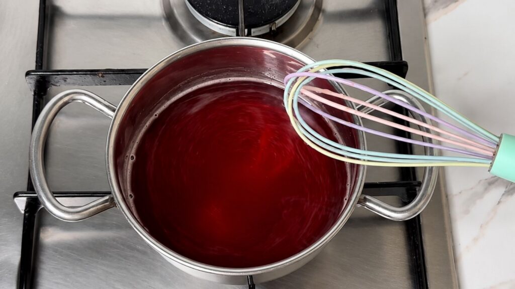 Preparing the strawberry cake glaze in a small saucepan by stirring water, sugar, and gelatin-based cake glaze mix until it just comes to a boil.