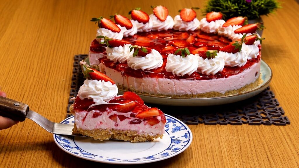 Close-up slice of no-bake strawberry yogurt cake on a blue-and-white plate, showing a crumb crust, creamy strawberry yogurt filling with fruit pieces, and a glossy strawberry topping with whipped cream and fresh strawberries in the background.
