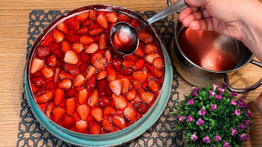 Piping the warm strawberry glaze over the sliced strawberries on top of the no-bake strawberry yogurt cake, using a large spoon to pour gently and evenly without melting the cream layer.