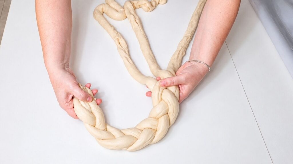 Braided dough being shaped into a Christmas bread wreath on a wooden work surface.