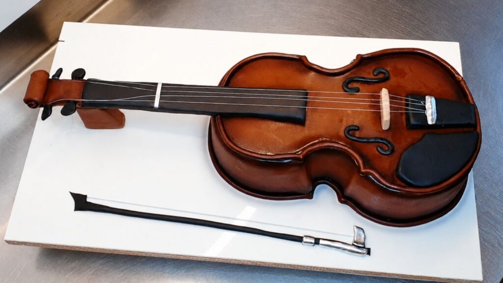Close-up of a realistic violin cake showing detailed fondant decorations, strings, bow, and airbrush shading for a lifelike finish.