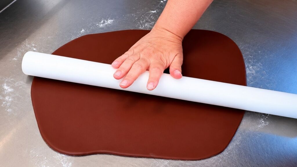 Rolling out brown fondant on a powdered sugar surface to cover a realistic 3D violin cake.