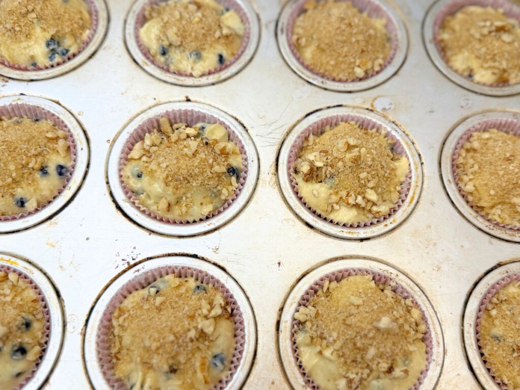 Blueberry muffin batter in a lined muffin pan topped with chopped walnuts and brown sugar