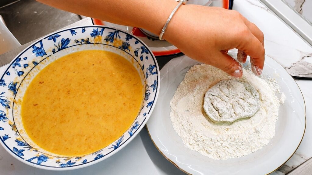 Coating an eggplant slice in flour before dipping it into the breading mixture.
