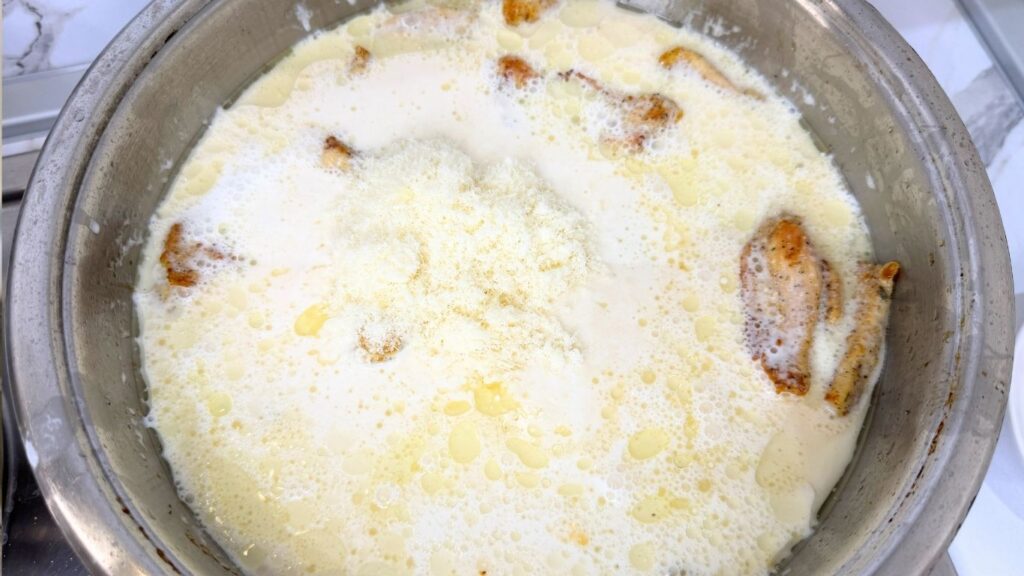 Adding cooking cream, Parmesan cheese, and water to the cooked chicken in a skillet.