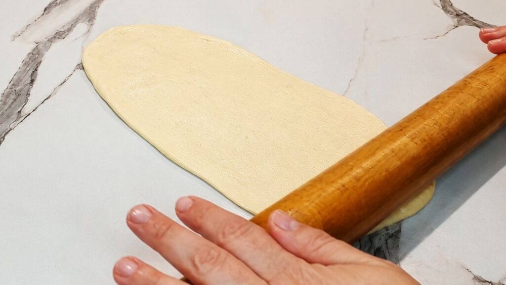 Rolling out a piece of soft dough with a wooden rolling pin on a lightly oiled work surface to shape a crescent roll.