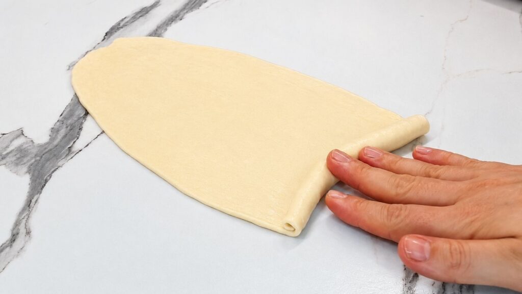 Using hands to gently roll a thin strip of dough from the wide end to the tip to shape a classic crescent roll.
