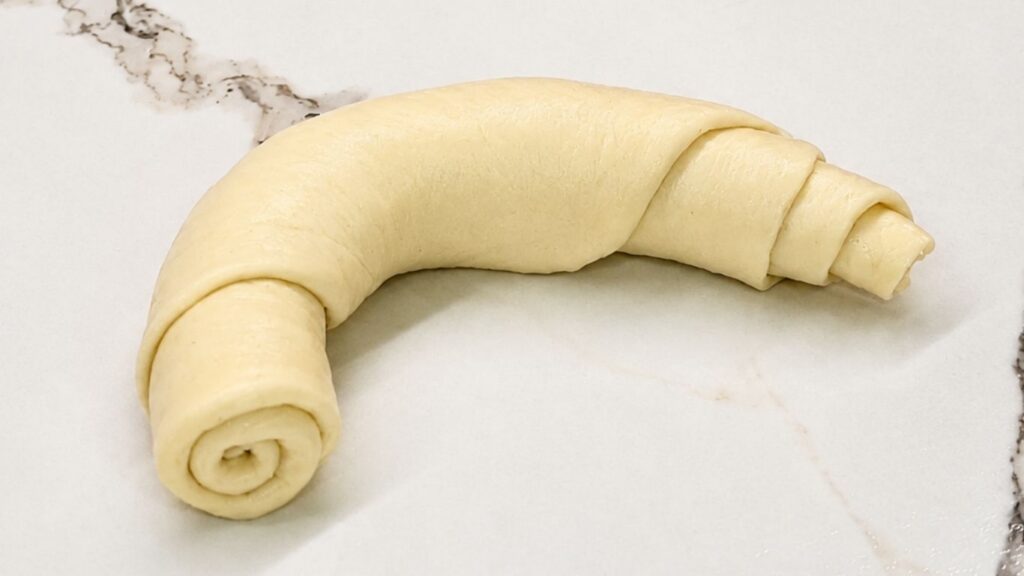 A fully rolled and shaped raw crescent roll resting on a work surface, showing its distinct layered spiral design.