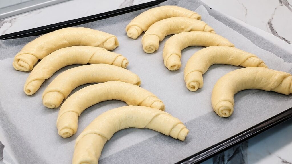 Shaped, raw crescent rolls arranged in rows on a baking sheet, ready for their final rise before baking.