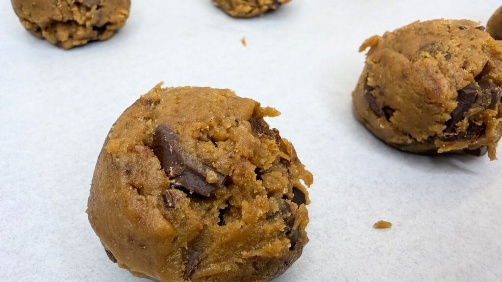 Scooped balls of chilled chocolate chip cookie dough arranged on a large baking sheet lined with parchment paper, ready to be baked in the oven.