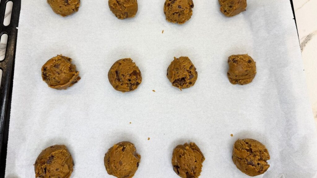 Scooped balls of chilled chocolate chip cookie dough arranged on a large baking sheet lined with parchment paper, ready to be baked in the oven.