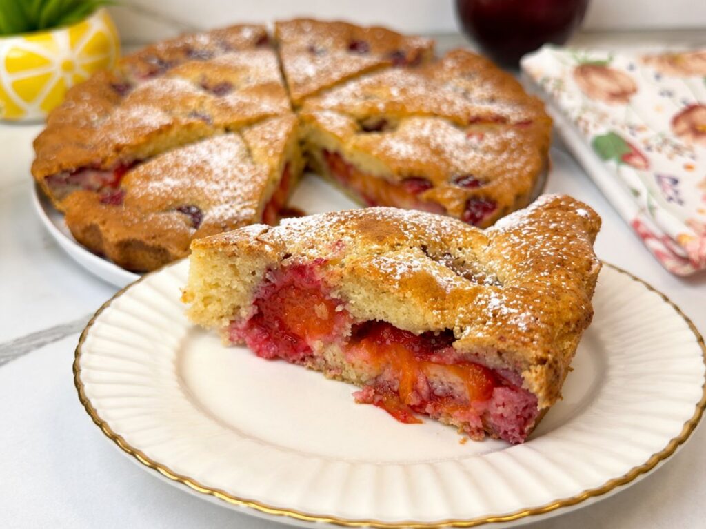 Slice of fresh plum cake with juicy plum pieces and powdered sugar on a blue-and-white plate.