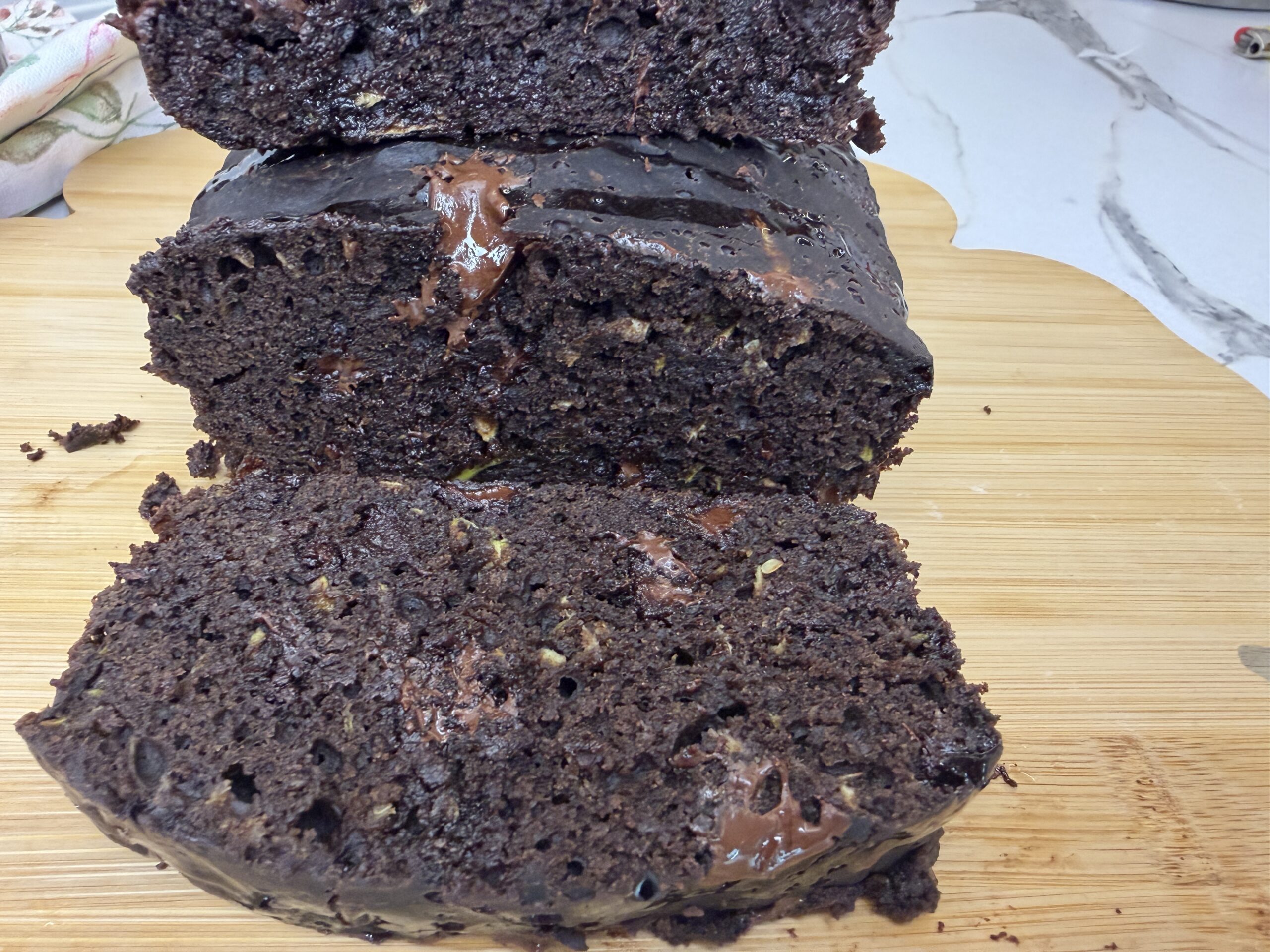 Moist chocolate zucchini bread sliced on a wooden cutting board with melted chocolate pieces on top.
