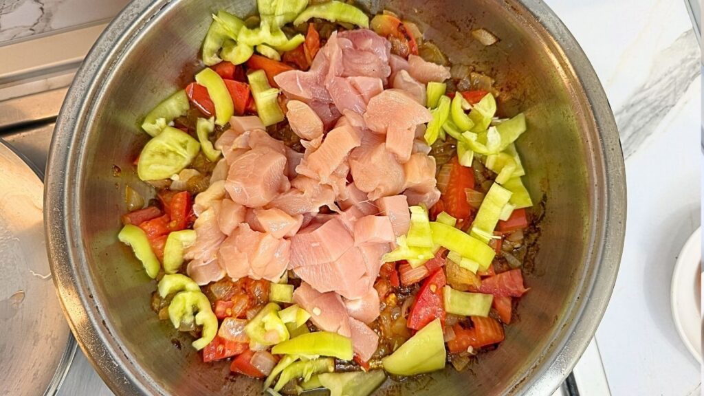 Adding chopped sweet peppers, tomato, and chicken pieces to the sautéed onion in a pot for one-pot chicken and rice.
