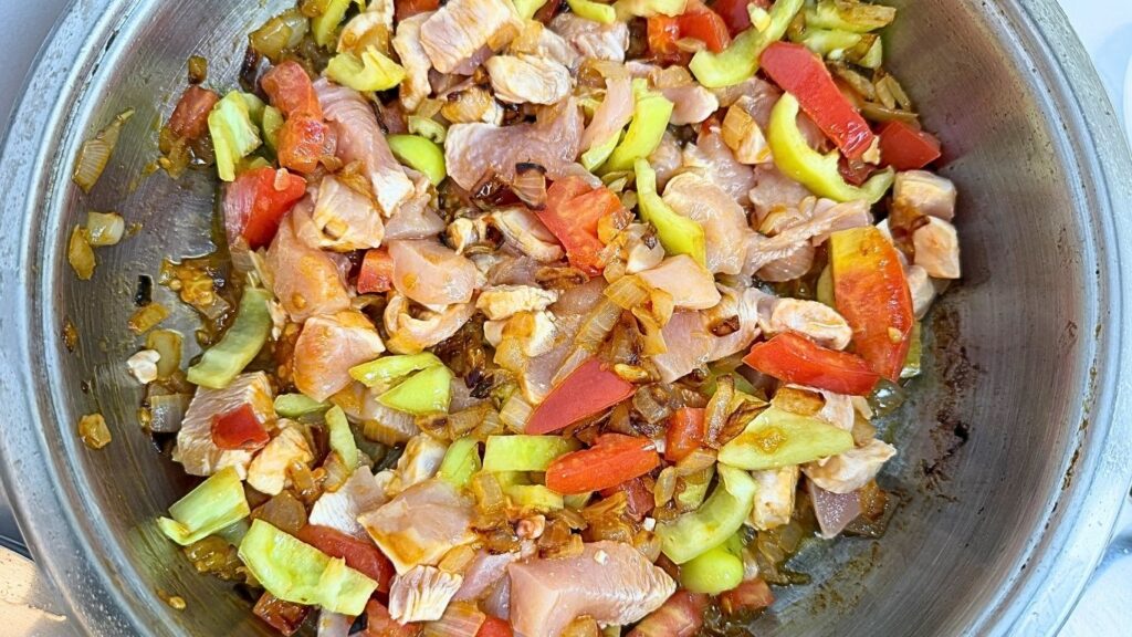 Adding chopped sweet peppers, tomato, and chicken pieces to the sautéed onion in a pot for one-pot chicken and rice.
