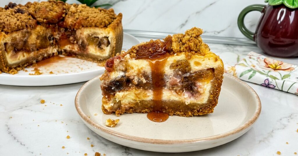 Slice of peach cobbler cheesecake with juicy peaches, buttery crumble topping, and sauce, with the whole cheesecake in the background.