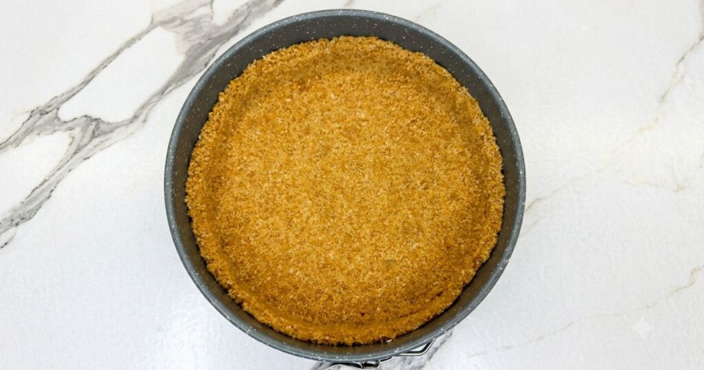 Graham cracker crust mixture pressed into a springform pan for Peach Cobbler Cheesecake.