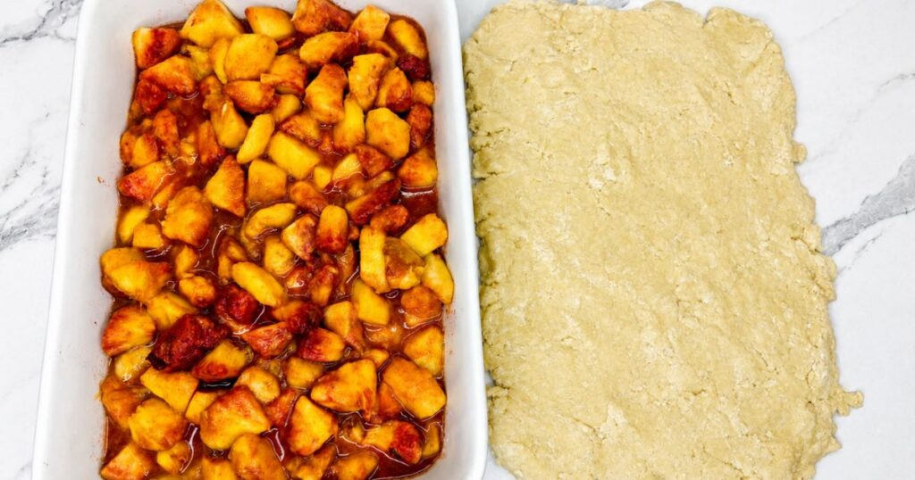 Baked peach filling in a baking dish with flattened cobbler topping pieces ready to be added on top.