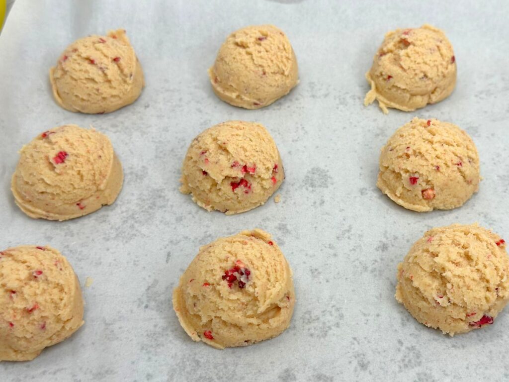 Scoops of raspberry sugar cookie dough arranged on a parchment-lined baking sheet, ready to bake.