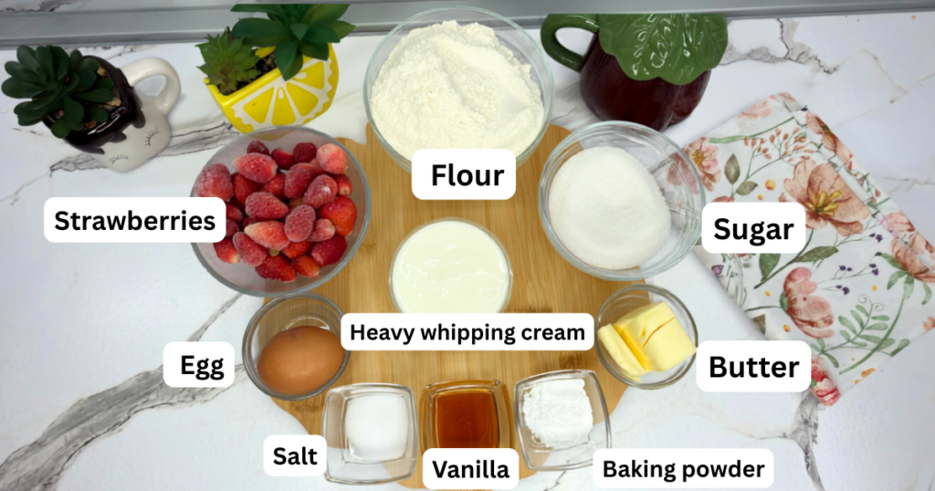 Ingredients for strawberry scones arranged on a table, including flour, sugar, butter, egg, cream, vanilla, and strawberries.