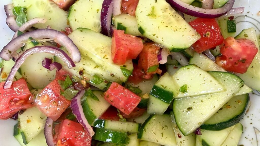 Pouring fresh lemon and olive oil dressing over chopped cucumber, tomato, and red onion for summer cucumber salad.