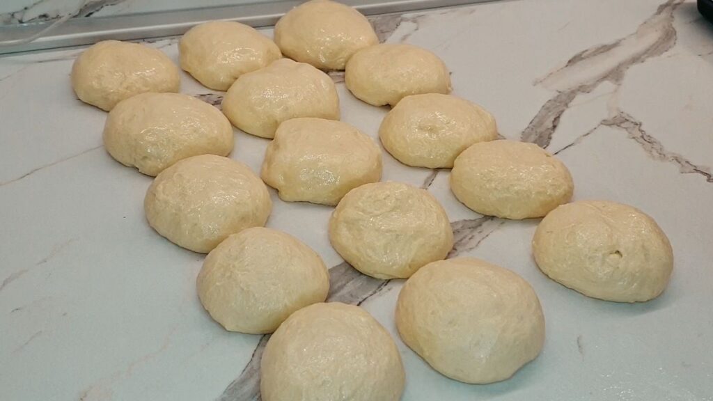 Soft crescent roll dough divided and shaped into 15 equal-sized dough balls on a lightly oiled wooden work surface.