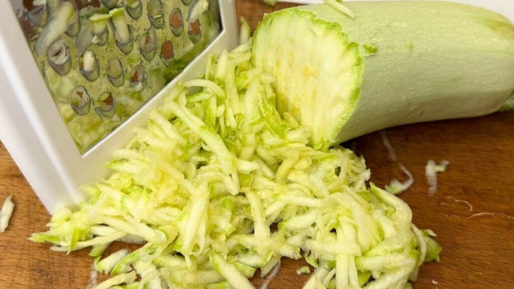 A close-up shot of freshly grating a green zucchini on a metal box grater into a large bowl to make Moist Chocolate Zucchini bread.