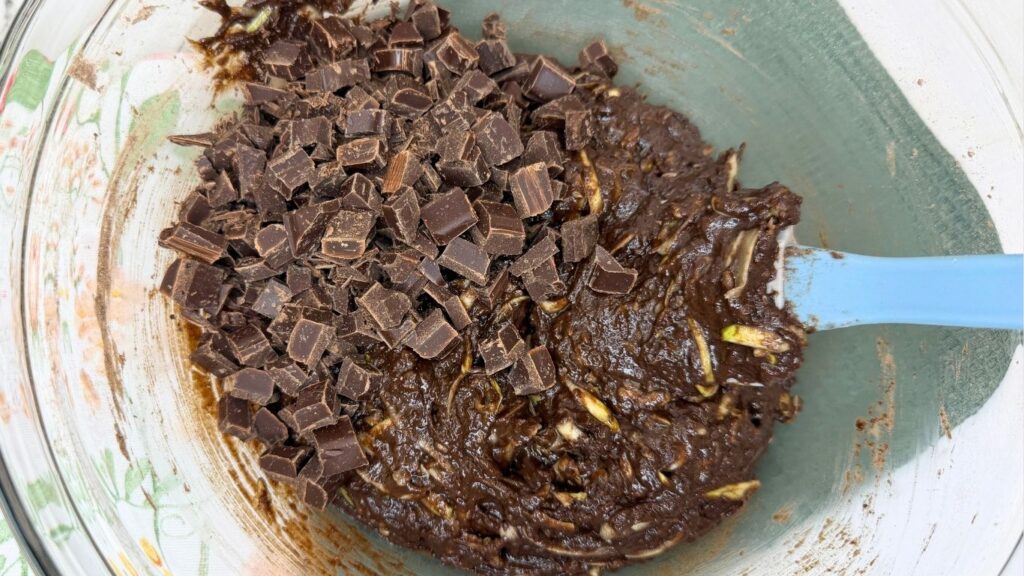 Folding dark chocolate chunks into a rich, thick batter in a glass bowl to make Moist Chocolate Zucchini bread.