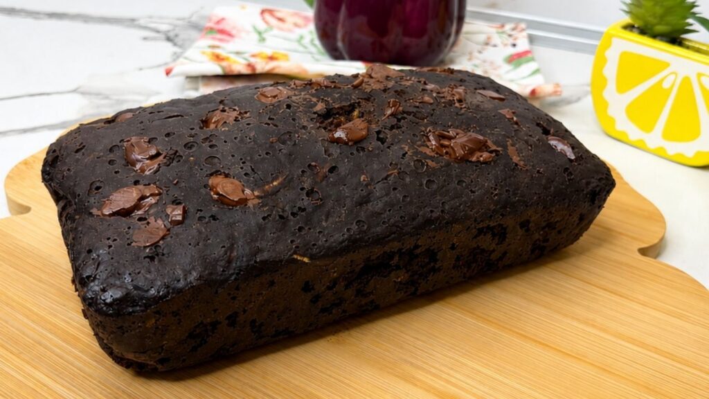 A freshly baked loaf of Moist Chocolate Zucchini bread cooling on a rustic wooden cutting board, showing its cracked, fudgy top and melted chocolate chips.