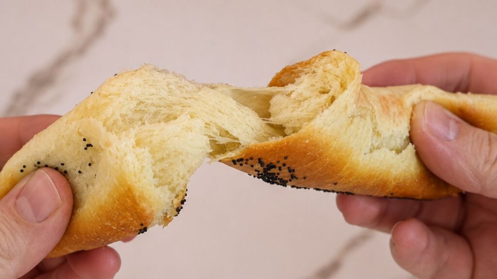 A close-up of hands holding and pulling apart a soft homemade crescent roll to reveal its fluffy, airy interior.