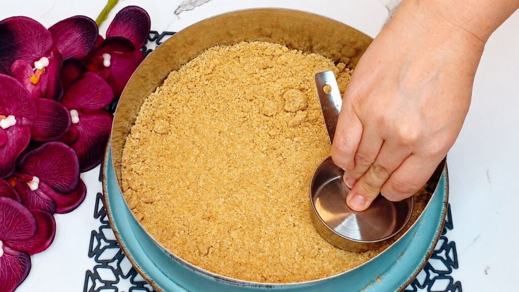 Crushed biscuit crust mixture evenly pressed into a cake pan for easy no-bake peach yogurt cake.