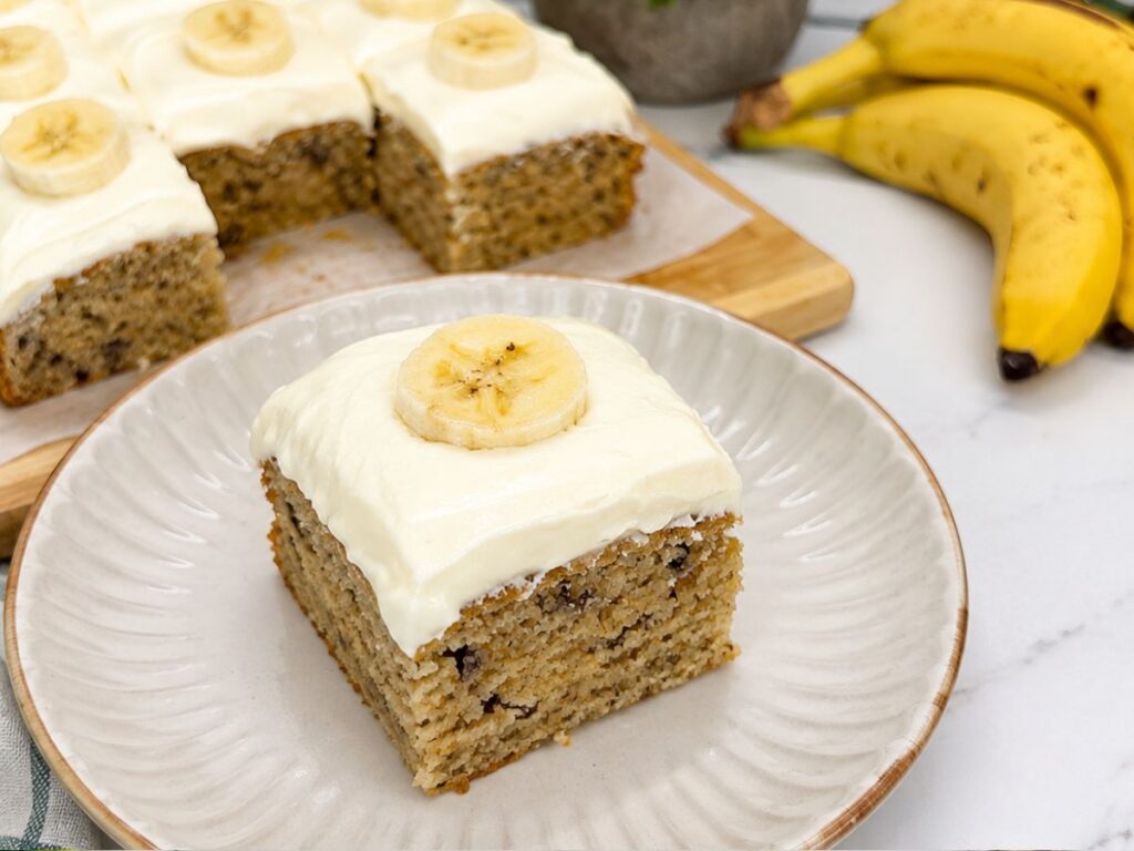 Sliced banana cake with cream cheese frosting and fresh banana slices in a baking pan.