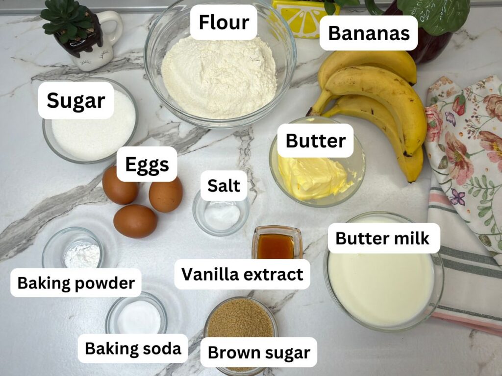 Banana cake ingredients arranged on a table, including ripe bananas, flour, butter, eggs, buttermilk, sugars, and cream cheese.
