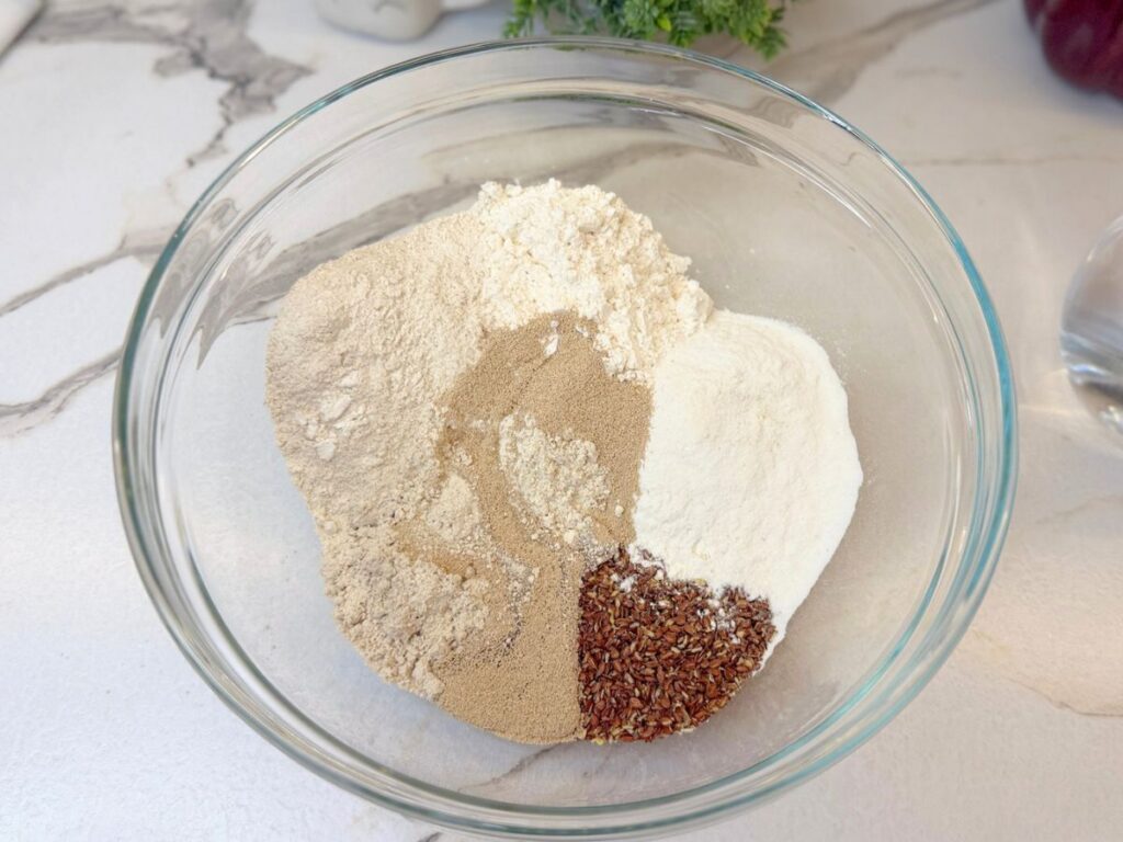 Gluten-free flours, yeast, salt, and flaxseed combined in a large glass mixing bowl.