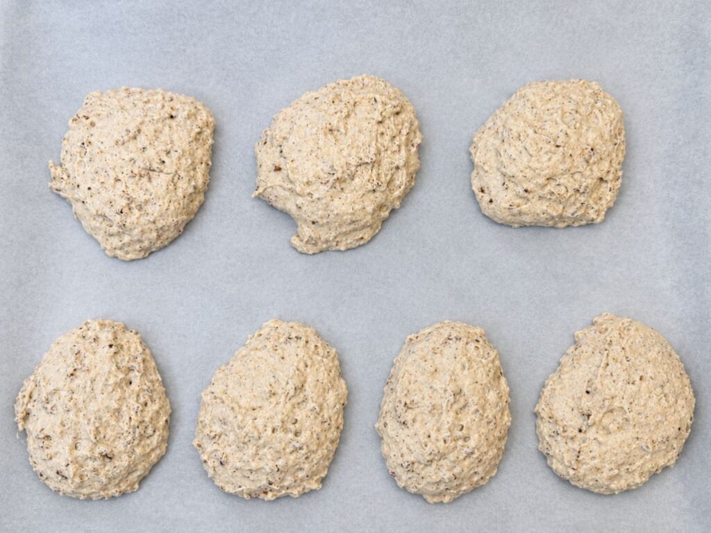 Seven portions of sticky gluten-free roll dough arranged on a parchment-lined baking sheet.