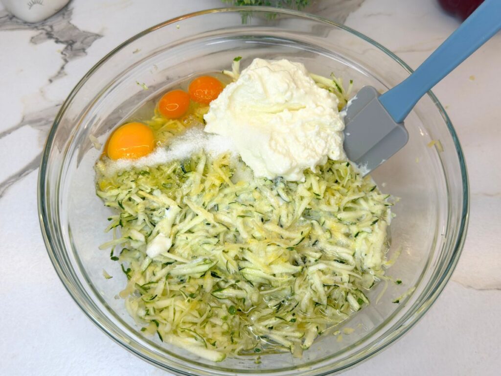 Grated zucchini with eggs, cottage cheese, buttermilk, oil, baking powder, and salt in a mixing bowl.