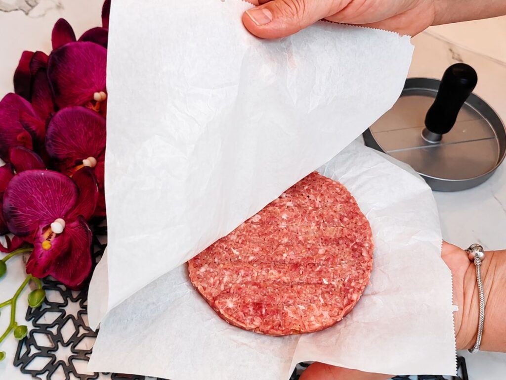 Raw burger patty shaped between parchment paper using a burger press