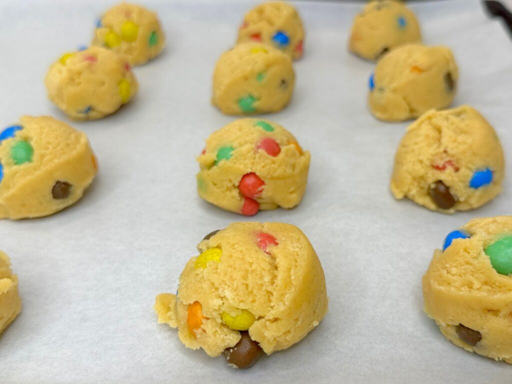 Shaped M&M cookie dough balls arranged on a parchment-lined baking sheet and ready to bake.