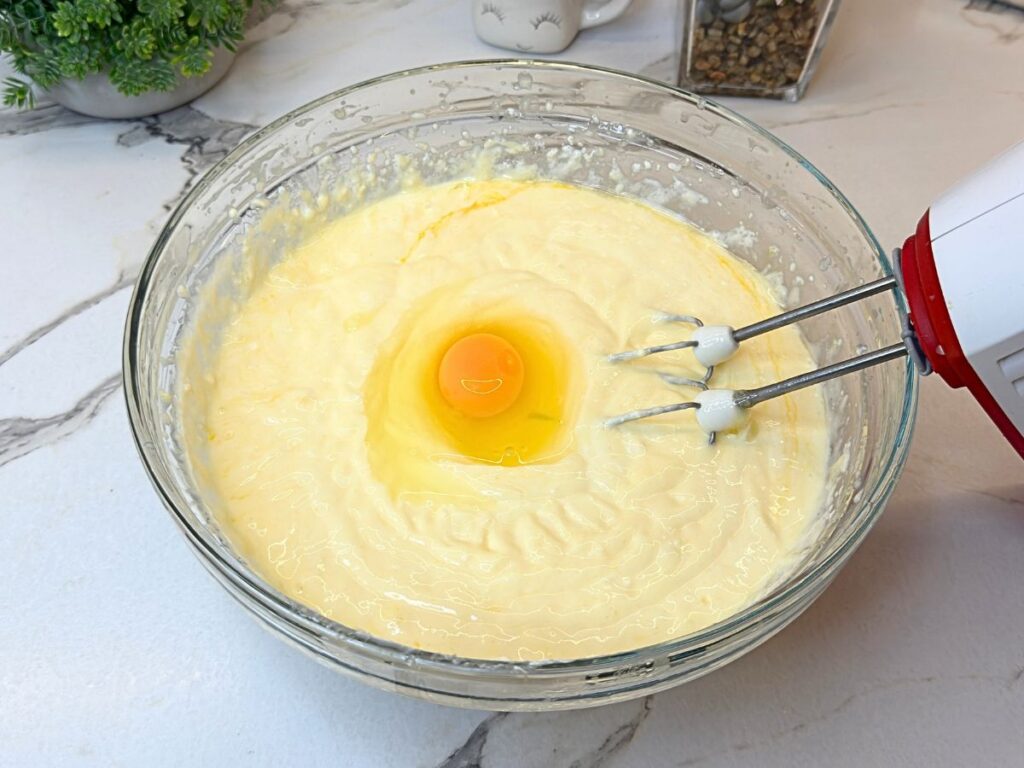 Egg being added to creamy New York cheesecake filling in a mixing bowl