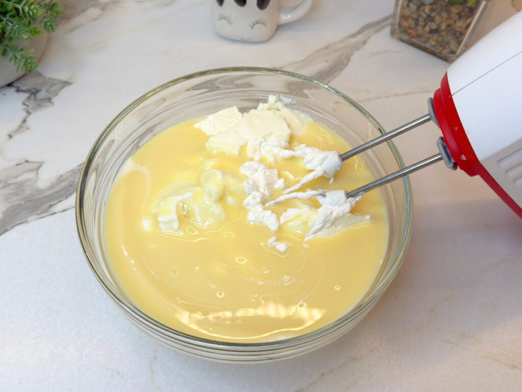 Cream cheese and sweetened condensed milk being mixed with an electric mixer.