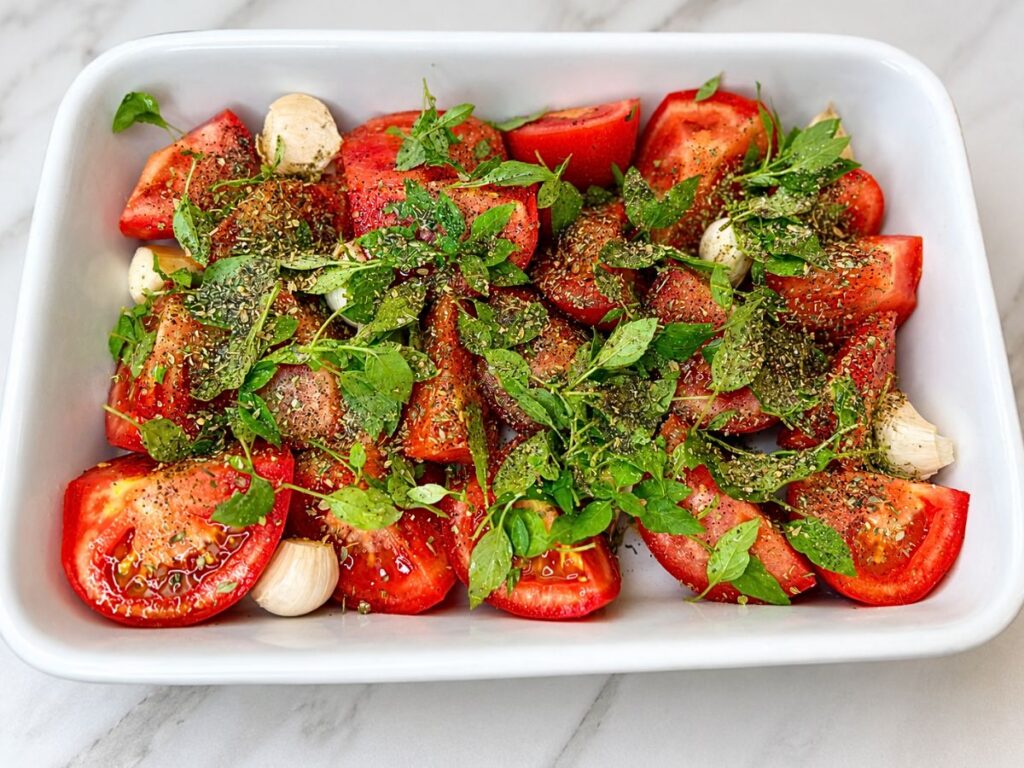 Quartered tomatoes and unpeeled garlic cloves seasoned with olive oil, oregano, and fresh basil in a baking dish