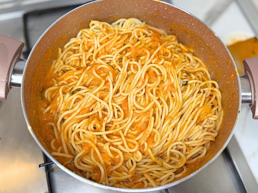 Spaghetti being tossed with smooth roasted tomato and garlic sauce in a large saucepan