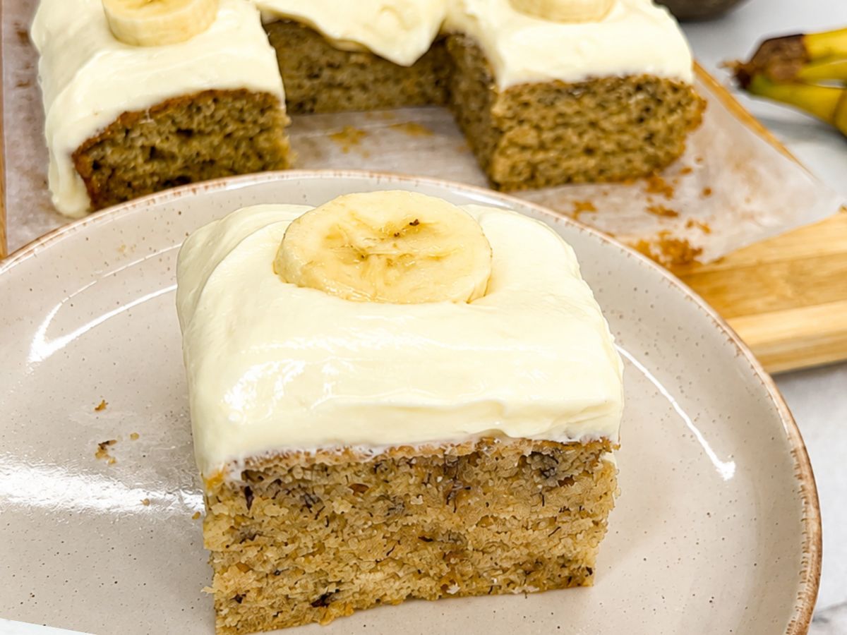 Moist banana cake slice with cream cheese frosting and fresh banana slices on top.