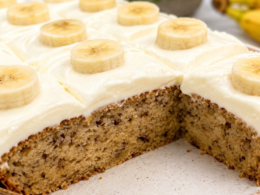 Sliced banana cake with cream cheese frosting and fresh banana slices in a baking pan.