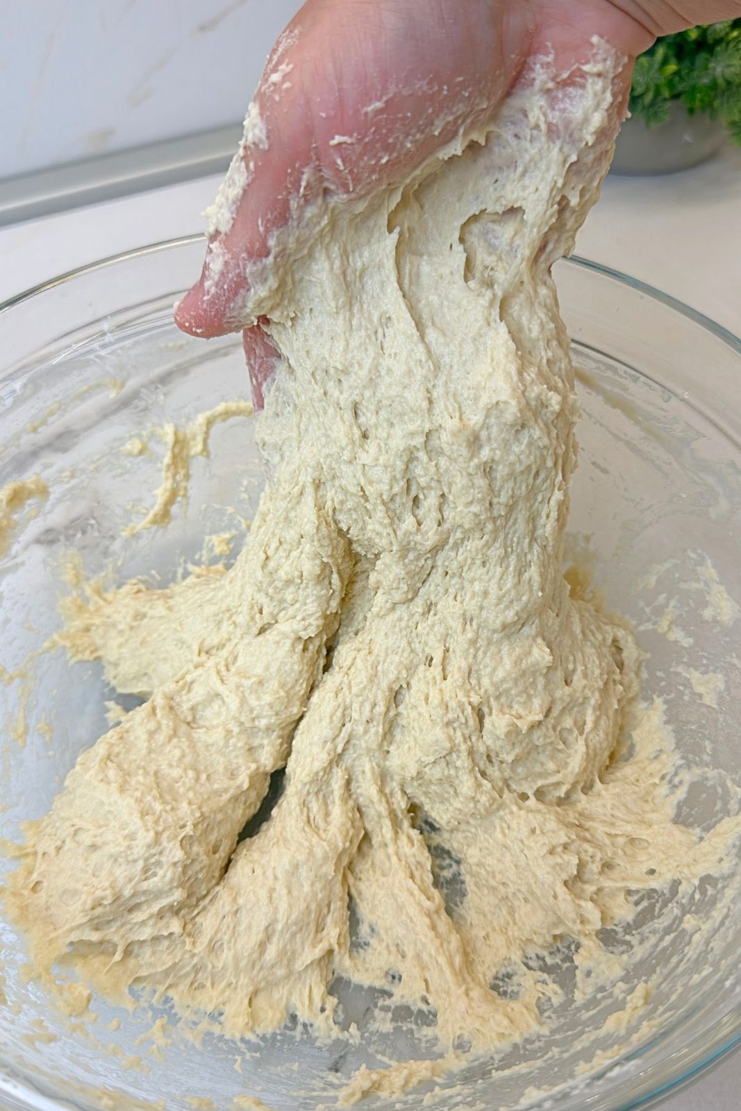 Soft sticky bread dough being gently kneaded by hand in a glass bowl
