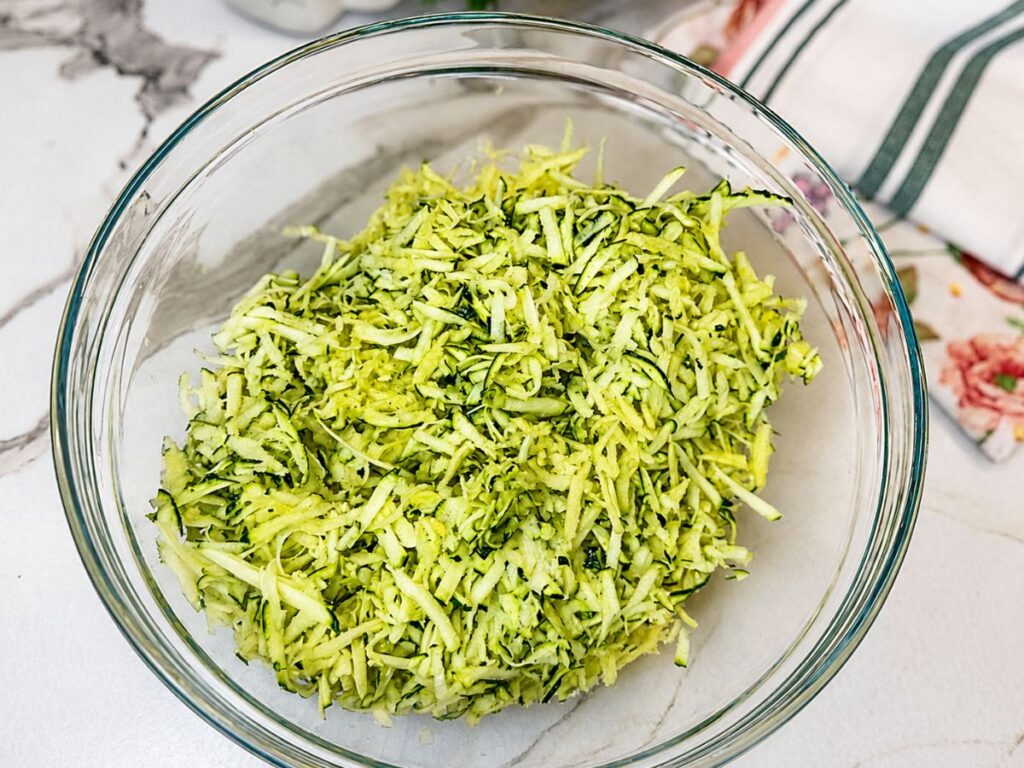 Freshly grated zucchini in a glass mixing bowl for making zucchini fritters.
