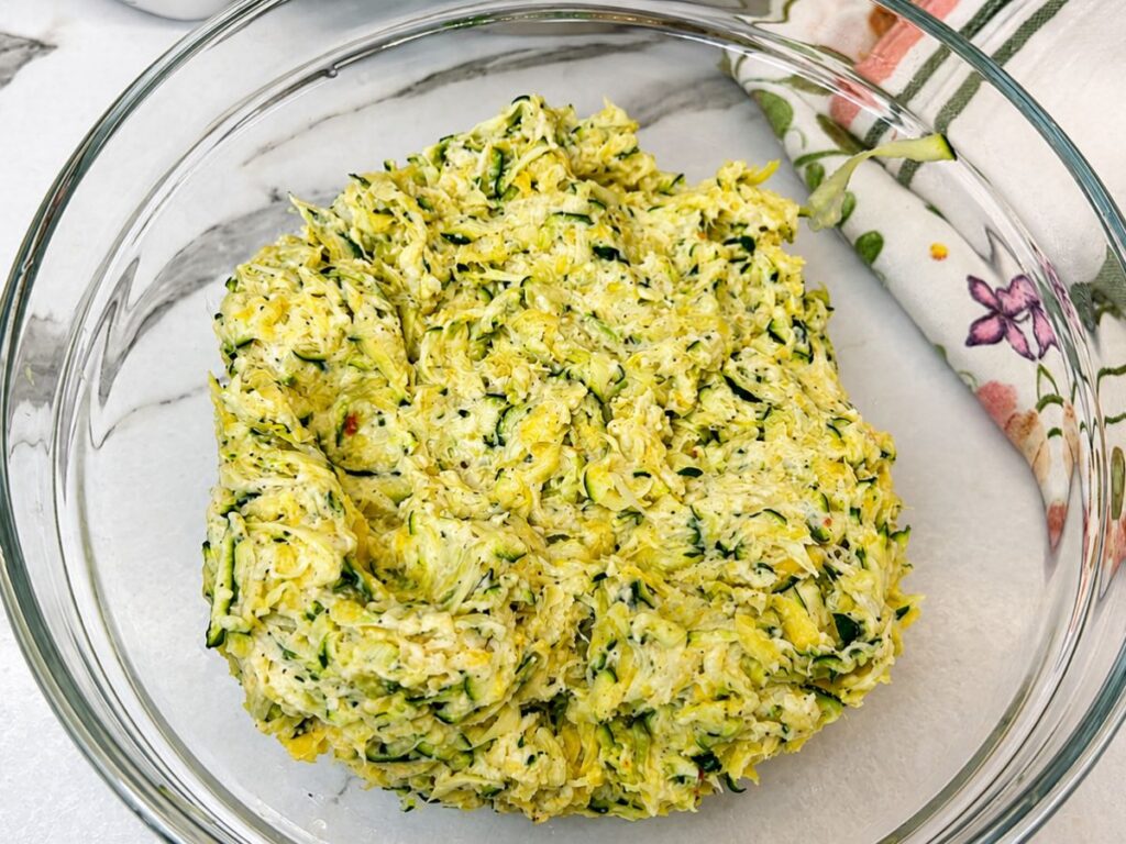 Zucchini fritter mixture combined with eggs, onion, flour, and seasonings in a glass bowl.
