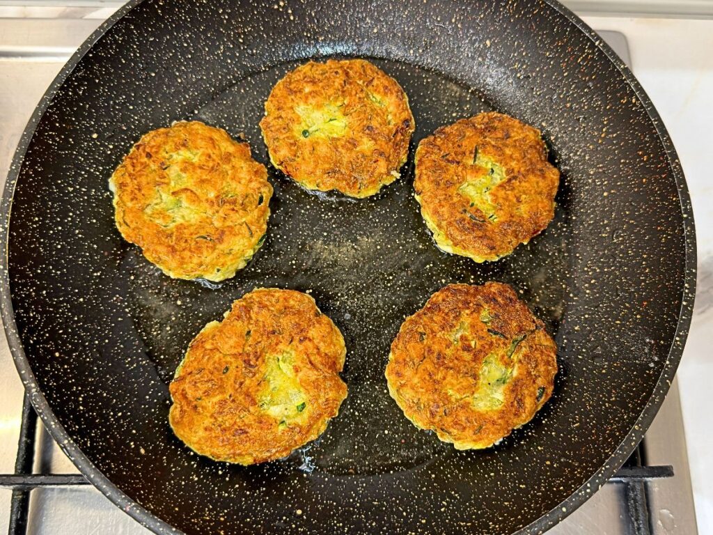 Crispy zucchini fritters frying in a skillet until golden brown on both sides.
