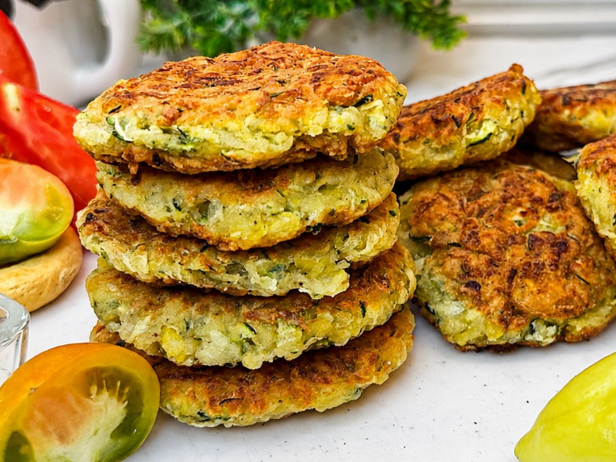 Easy crispy zucchini fritters stacked on a plate and served with sour cream, cucumber slices, fresh herbs, and lemon wedges.