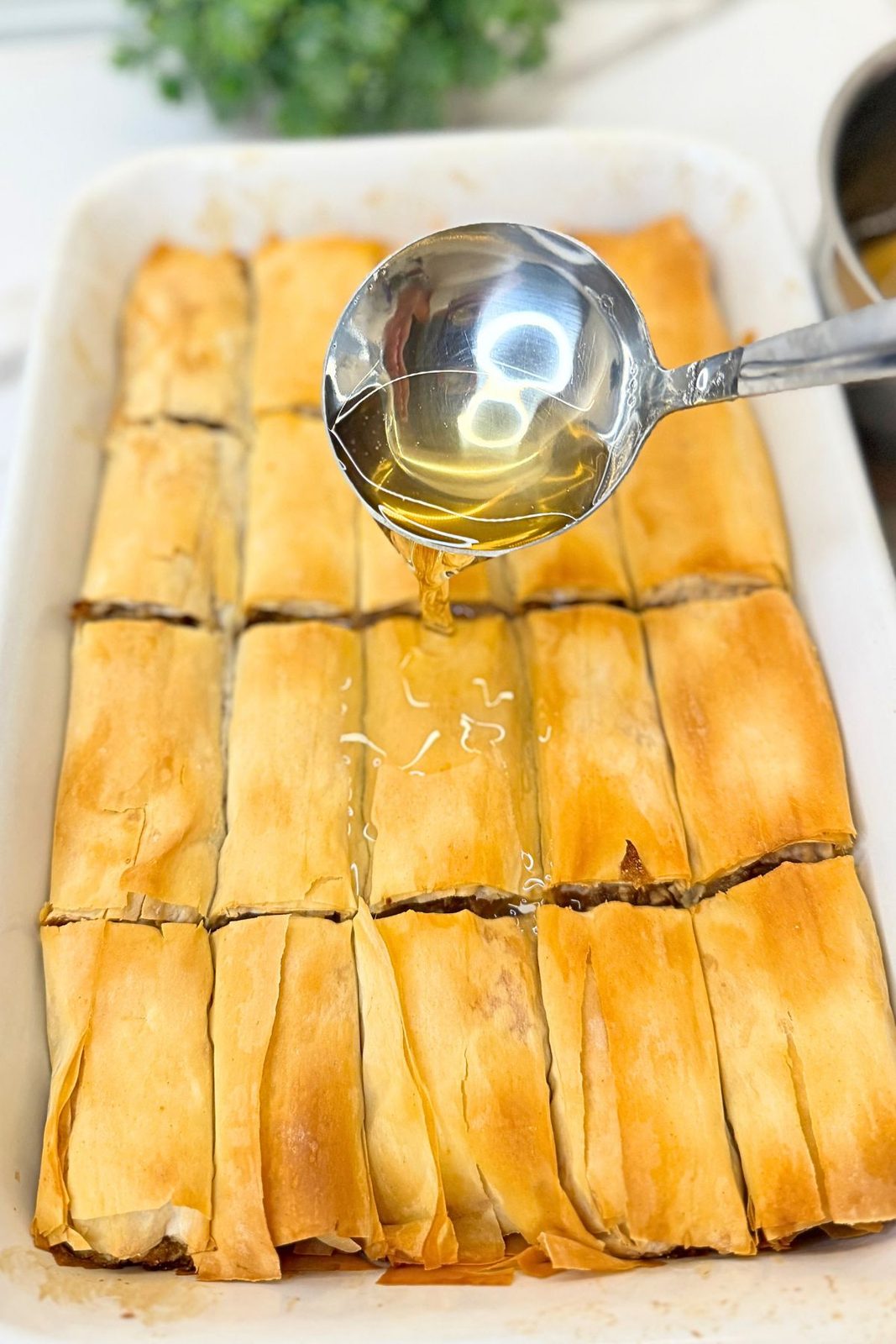 : Hot vanilla syrup poured from a ladle over golden baked apple phyllo rolls in a white baking dish.